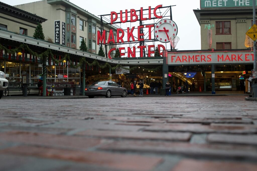 xterior view of Pike Place Market in Seattle, featuring the iconic red neon sign and clock, with people walking nearby and the historic building in the background.